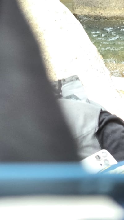 The image shows a partial view of what appears to be a person standing near a body of water or riverbank. The person is wearing a black top and grey trousers or shorts. There is a light-colored, possibly concrete, ledge or structure to the left, and water is visible beyond it. The presence of a phone suggests a contemporary setting. There is not enough information to place this scene in a location in Nam Ninh Hoa, Vietnam.