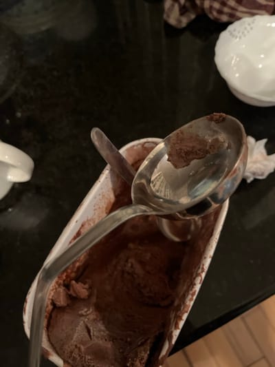 The scene depicts a partially consumed rectangular white cardboard container of chocolate ice cream situated on a dark, reflective countertop in a kitchen in Joliet, United States of America. Inside the container, a large, reflective silver metal ice cream scoop rests with its bowl facing upwards, containing a small amount of chocolate ice cream residue. A smaller, dark-colored spoon is also present within the container, partially submerged in the ice cream. The chocolate ice cream shows signs of having been scooped, with an uneven, slightly softened surface. In the upper right background, a white ceramic bowl with a textured rim is visible, along with a brown and white checkered cloth. A partial view of another white ceramic object is present in the upper left background. No individuals are visible.