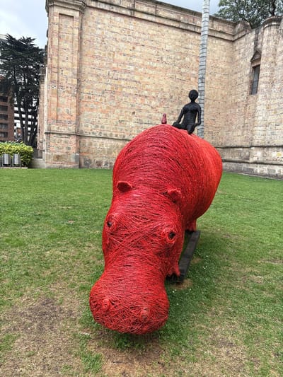 A red, life-sized hippo sculpture made of interwoven material sits on green grass in front of a brick building. A smaller, black statue of a person is on the hippo's back. A palm tree is visible to the left of the hippo, and buildings can be seen in the background. The ground is covered in grass, and a dark object is sticking out behind the hippo.