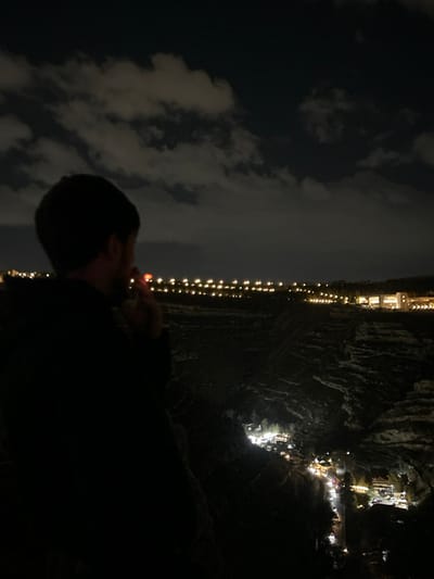 The image captures a nighttime panoramic view from an elevated position within Dummar Municipality, Syria. In the immediate left foreground, the silhouette of a male figure is visible from the back and right side. He possesses short dark hair and a beard, and wears a dark outer garment. A small, distinct orange glow is present near his mouth, indicating he is holding a lit object, consistent with a cigarette. The individual is oriented towards the expansive cityscape and natural landscape.

The scene encompasses a deep, dark valley or gorge. Along the base and lower slopes of this valley, a winding path or road is illuminated by a series of bright lights from vehicles, streetlights, and buildings or establishments. In the mid-distance, a long, horizontal line of uniformly spaced bright lights extends across the terrain, suggesting a significant roadway or infrastructure at a higher elevation. Further into the background, several illuminated multi-story buildings mark a distinct urban area on the horizon. The sky overhead is dark, with scattered, irregularly shaped clouds visible.