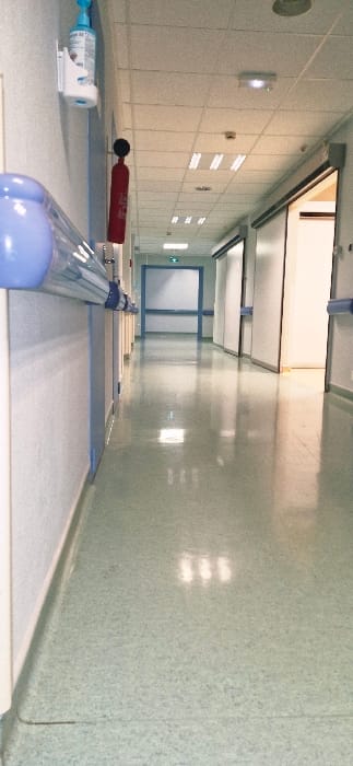 A sterile, tiled hallway extends into the distance with a high ceiling featuring evenly spaced light fixtures. On the left are tiled walls with integrated lights. A blue pipe runs along a ledge with various medical items scattered nearby, including a red object and a bottle. At the end of the hallway is a set of closed, frosted glass doors with a green light above them. The room has a clinical and utilitarian feel.