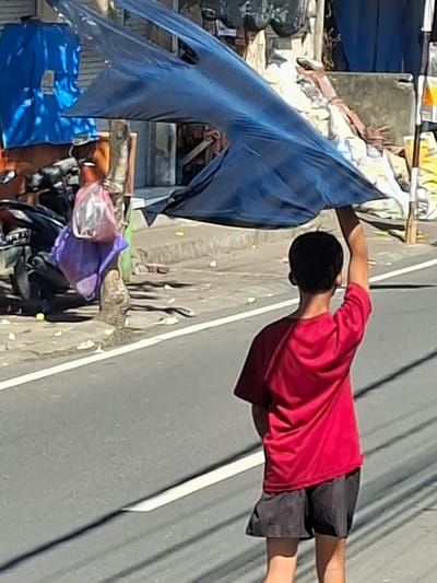 In Blahbatuh, Indonesia, a young person wearing a red shirt and black shorts walks on a street, holding a large, dark blue tarp above their head as a makeshift umbrella. The tarp is distorted by the wind, creating an abstract shape. Behind the person, a motorcycle is parked near a tree with bags, including a purple one and a clear plastic bag containing items, hanging from it. The environment suggests a street scene with some buildings and structures in the background, characterized by a somewhat cluttered and outdoor setting. The road has white markings, indicating it is a paved surface. The lighting suggests it is daytime.
