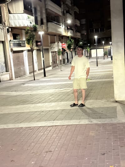 A young man wearing a light-colored t-shirt and light-colored shorts with black slides walks across a pedestrian crossing at night. The pedestrian crossing is made of gray and white striped paving stones. The man is in focus and is walking towards the right of the frame. The background consists of a street scene with buildings, streetlights, and closed storefronts, suggesting a commercial area in Alicante, Spain. The overall lighting is artificial, likely from streetlights and building lights, casting shadows on the ground.