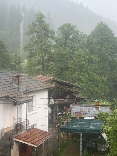 The image captures a residential area in Elhovets, Bulgaria, during heavy rainfall, evidenced by visible rain streaks throughout the scene. The background features densely forested hills and mountains, largely obscured by mist and rain. A waterfall is visible cascading down a distant, tree-covered slope on the upper left.

In the mid-ground, on the left, a light-colored, two-story house with a dark, ridged tiled roof is prominent. A brick chimney, showing signs of weathering and missing bricks, protrudes from this roof, secured with metal flashing. A white satellite dish is affixed to the side of the building. An exterior staircase descends to a ground-level porch featuring a wrought-iron railing and a terracotta-tiled awning. Multiple electrical wires and cables traverse the scene, connecting to the buildings.

To the right of the main house, a cluster of smaller, utilitarian structures is visible. One structure has a partially exposed or unfinished wooden roof frame, with its interior contents partially visible. Adjacent to it, another structure features a heavily rusted corrugated metal roof, from which a smaller brick chimney emerges. A dark green tarp is stretched over an outdoor area located between these structures, possibly covering furniture or equipment. The immediate foreground and areas surrounding the structures are rich with green foliage, including various trees and bushes. No human or animal figures are visible in the scene.