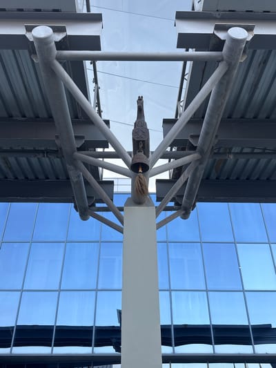 The image depicts an architectural detail, likely an exterior element of a building, with a prominent metal structure supporting a roof or overhang. The metalwork is composed of grey, cylindrical beams and supports, some appearing weathered. Suspended from the structure is a decorative bell with a tassel, and above it, a carved object resembling an animal head, possibly a horse. The background consists of a large, blue-tinted glass facade, reflecting the sky and surrounding environment. The perspective is from below, looking upwards at the architectural features. The scene is set in Yakutsk, Russia, suggesting a contemporary urban setting. There are no people or immediate actions depicted in the image. The overall impression is one of architectural design and decoration.