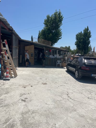 In Laneia, Cyprus, a black hatchback car is parked on a cracked concrete lot outside a cluttered open-air workshop. The workshop has a corrugated metal roof and shelves displaying various items including chains, rolls of material, and tools. Two men are visible within the workshop. One man, wearing a grey t-shirt and shorts, stands near the entrance. Another man, in a dark t-shirt and camouflage shorts, is further inside. To the left of the workshop, a wooden ladder is leaning against a metal structure, with an orange and white traffic cone at its base. Tall green trees frame the scene under a clear blue sky.