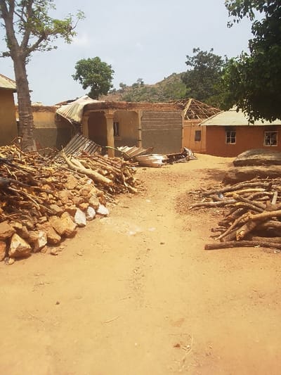 The scene is a dirt path in front of a house in Kwal, Nigeria. A large pile of rubble and branches is in the left foreground, followed by a dirt path leading toward a house in the background. The house is made of cement block and has a collapsed roof. A second house is visible in the background to the right, constructed of mud brick. The sky is partly cloudy.