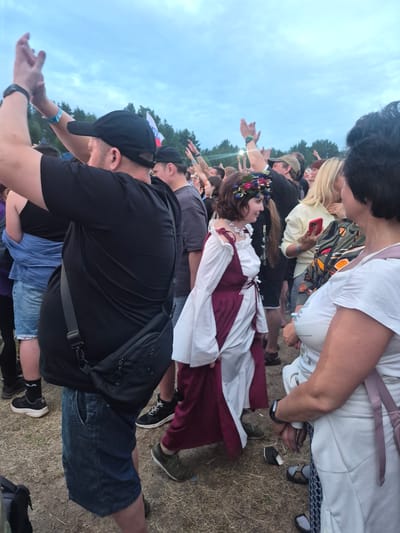 The image depicts a crowd of people at an outdoor event, possibly a music festival, given the raised hands and festive atmosphere. The sky is overcast with some clouds visible. In the foreground, a person wearing a black t-shirt and a black baseball cap is visible from the back, with their hands raised. To their right, a person in a white and burgundy medieval-style dress with a floral wreath on their head is facing left and smiling. Further to the right, a person with dark hair is holding a red smartphone. Another person with blonde hair is partially visible behind them. The ground appears to be covered in dry grass and dirt. A flag with white, blue, and red colors is visible in the background, possibly the Russian flag. The setting appears to be an open field surrounded by trees, consistent with a location outside a town or city. There is no specific visual evidence to confirm the location as Lagunovo, Russia.