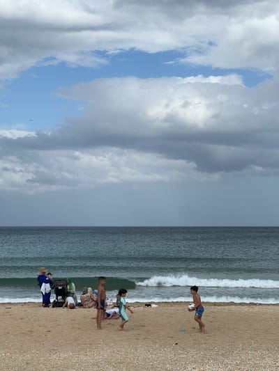 The image shows a beach scene in M'Diq, Morocco. The sky is partly cloudy. The sea is adjacent to the beach, with waves visible. A group of people is on the beach. One boy is running. Another boy with a ball. Others are standing, sitting, or pushing a stroller. The beach is sandy with some shell fragments.