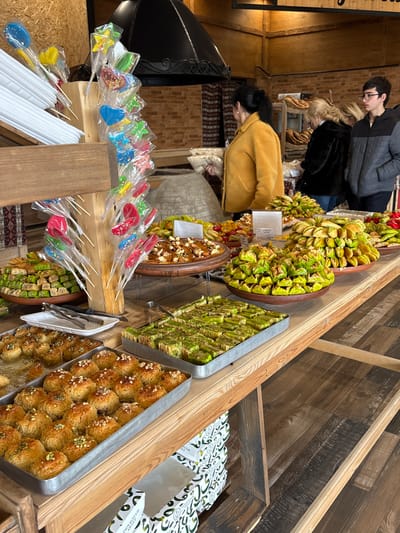 An indoor scene in Tsovagyugh, Armenia, depicts a retail establishment with a prominent display of various traditional sweets and pastries on a long wooden table. In the immediate foreground, to the left, a vertical wooden rack holds numerous colorful lollipops, individually wrapped in clear plastic, some shaped like hearts, stars, or ovals.
The wooden table displays a wide array of confectionery. On the left, a rectangular metal tray contains small, golden-brown pastries, possibly pakhlava or similar, topped with chopped nuts. Adjacent to this, a white plate with silver tongs is available for serving. To the right, another rectangular metal tray holds numerous green, pistachio-dusted rectangular pastries. Further along the table, several elevated round wooden platters are laden with different sweets: one with neatly stacked green, triangular-shaped pastries; another with a mixture of dried fruits and nuts; and additional platters featuring similar arrangements of green pastries and other confectioneries. Small white blank signs are interspersed among the sweets, likely for product identification or pricing. Below the table, stacked white cardboard boxes with green designs are visible, presumably for packaging.
In the mid-ground, three individuals are present near the display. A woman in a mustard yellow, long-sleeved jacket is positioned with her back mostly to the viewer, facing right, holding a white rectangular object, possibly a flatbread or packaging, near a traditional clay oven (tonir) situated behind the display table. To her right, another woman, wearing a black, textured jacket, also stands facing right. To the far right, a young male wearing glasses and a grey and black puffer jacket stands, looking towards the right side of the frame.
The background features an ornate black metal exhaust hood above the clay oven. The wall behind the oven is paneled with light wood, transitioning to exposed light brown brick further right. Behind the individuals, shelves stocked with various bread loaves are visible against a light-colored wall. The floor consists of dark wooden planks.