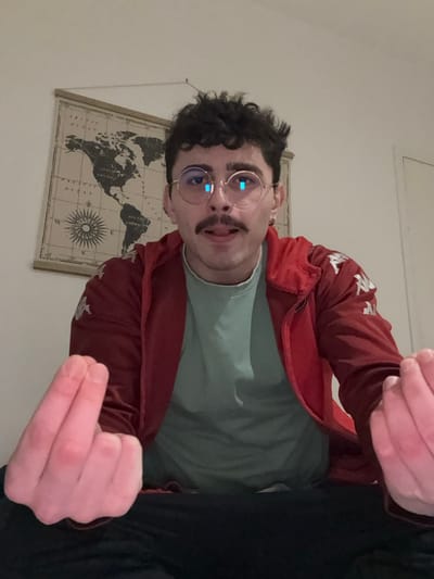 A person with curly dark hair and a mustache, wearing round glasses, a light green t-shirt, and a red Kappa jacket, is shown from a tilted, low-angle perspective. They are holding up two fingers in a gesture. A world map poster hangs on the wall behind them. Part of their black pants are visible on the right. The room's wall is painted white. The overall lighting in the image appears somewhat dim.