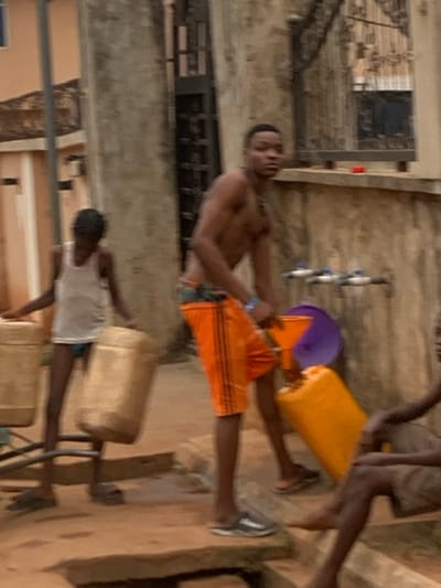Several people are filling plastic containers with water from a public tap. One shirtless man in orange shorts stands at the tap, holding a yellow container. Behind him, someone holds a purple basin. A younger person in a white shirt fills a tan container on the ground. The scene takes place outdoors, with buildings visible in the background. The image is slightly blurred.