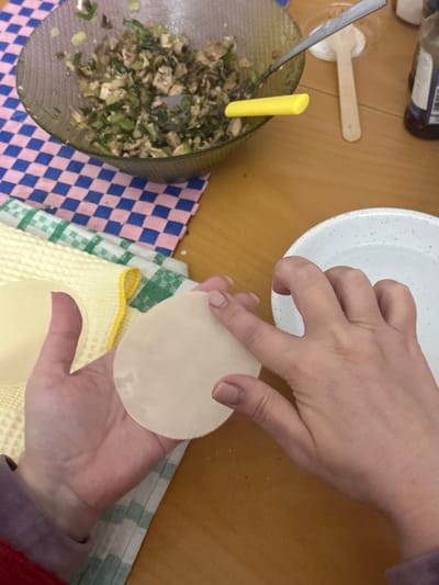 The image provides a top-down view of a person's hands engaged in the process of preparing dumplings on a light brown table or kitchen counter, suggesting an indoor home cooking environment.

The main subjects are a pair of hands, likely a woman's, holding two round, thin dumpling wrappers. The left hand supports one wrapper while the right hand holds another, positioned as if to begin filling or folding. The hands exhibit light-colored nail polish.

To the left, a clear or mesh bowl contains a generous amount of dumpling filling, composed of finely chopped green vegetables and lighter ingredients, possibly meat or tofu. A yellow-handled spoon and a metallic spoon are partially submerged in the filling, indicating they are in use. Further left on the table, part of a dark bottle and a wooden spoon are visible.

In the upper center, a white speckled bowl is present, likely intended for water to seal the dumplings or to hold more wrappers. The table is adorned with several patterned textiles: a blue and pink checkered cloth on the lower left, and a green and white checkered cloth along with a yellow waffle-weave towel beneath the hands and wrappers, providing a clean workspace.

The activity is clearly the meticulous assembly of dumplings, with all components laid out for the task. No specific time of day or weather can be discerned from the indoor setting, and no textual information is visible on any objects. The scene depicts a calm and focused cooking activity taking place in Egkomi Lefkosias, Cyprus.