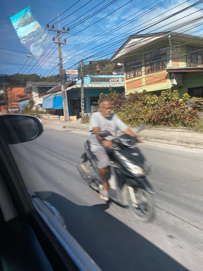 A man is riding a scooter on a street in Baan Hua Thanon, Thailand. He is wearing a light-colored t-shirt and shorts, and white flip-flops. The scooter is black. The man is in motion, and the background is blurred. The background consists of buildings, trees, and a blue sky with some clouds. There are power lines visible across the sky. The image appears to have been taken from inside a vehicle.