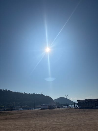 The image depicts a wide, sun-drenched landscape in Turka, Russia, with a clear blue sky dominating the upper portion. A bright sun with prominent rays and a lens flare is positioned in the upper center of the frame. Below the sky, a line of dark green, densely forested hills stretches across the horizon, with a single transmission tower visible on one of the peaks. In the foreground, a vast expanse of dry, sandy ground shows tire tracks, suggesting it is a road or a parking area. Along the shoreline, a body of water is visible, with a few boats moored and a walkway or pier extending into the water. Several buildings, including a small kiosk labeled "КАССА" (KASSA - meaning "cash desk" or "ticket office" in Russian) and a larger structure with signs in Russian, are present along the waterfront. A dark-colored SUV is parked on the sandy ground to the left of the kiosk, and a small tractor-like vehicle is visible near the water's edge. The overall scene conveys a sense of a sunny day in a waterfront area with some commercial activity.
