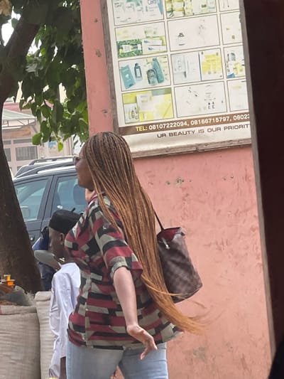 The image captures an outdoor street scene in Samaru, Nigeria, during daylight. A woman with dark skin is prominently featured in the mid-ground, walking from right to left. She wears eyeglasses, a patterned short-sleeved top with red, black, and greenish-gray blocks, and light blue jeans. Her long, light brown braided hair extends below her waist, appearing to be in motion. She carries a brown checkered shoulder bag with a red interior lining on her left shoulder. Her head is turned to the left, revealing a smiling or laughing expression. Behind her and to the left, partially obscured by a tree trunk, stands a man with dark skin wearing a black skullcap and a white shirt. To the right, a faded pink wall displays a white rectangular advertisement sign for beauty products, featuring images of items like CeraVe, Nigerian phone numbers, and the slogan "UR BEAUTY IS OUR PRIORITY." In the left background, a dark-colored SUV is visible, and below the tree, white sacks and small containers with amber liquid indicate a street vendor's display.