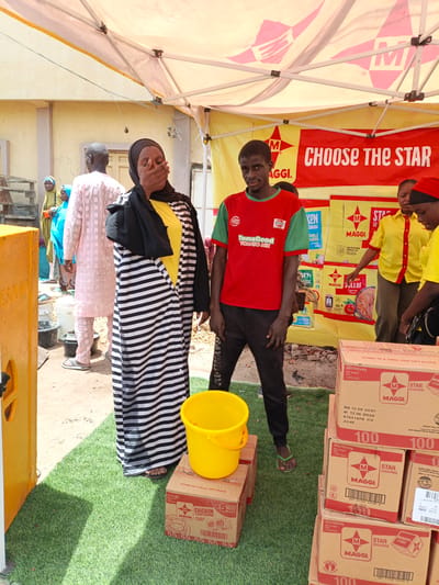 In Tarauni, Nigeria, a promotional event is taking place under a white tent with yellow trim. Several people are present. In the foreground, a woman wearing a black hijab and a black and white striped robe, with a yellow shirt underneath, is covering her mouth with her hand. To her right, a man in a red and green t-shirt and black pants stands looking towards the camera. Behind them, more people are visible, including a man in a pink patterned tunic and trousers, and others in yellow shirts. A red banner with the text "CHOOSE THE STAR" is displayed behind the individuals. Several boxes labeled "MAGGI STAR SEASONING" are stacked on artificial green grass in the foreground and to the right. A yellow bucket is placed on top of one of the boxes. The environment appears to be an outdoor market or street setting.