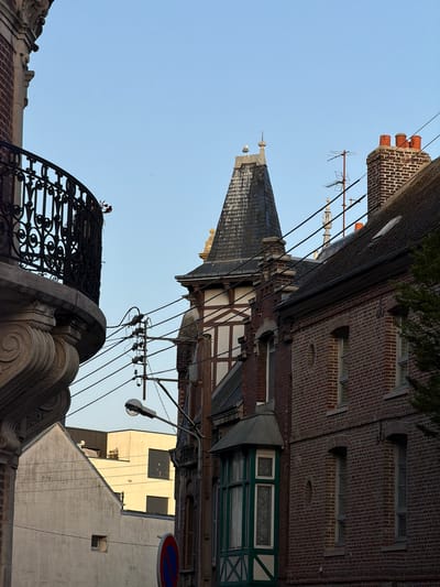 This image shows a view of buildings and architecture in Dieppe, France. The perspective is from below, looking up at rooftops and facades against a clear blue sky.
The main subjects are architectural elements of various buildings. We see a steeply pitched, dark-tiled roof of a historic building, possibly with dormer windows and a spire. Adjacent to this is a brick building with exposed timber framing in a half-timbered style. There are several windows visible, some with decorative green frames and patterned glass. Another brick building is visible with multiple windows and ornate brickwork around them. Further in the background, a lighter-colored building with multiple rectangular windows is present.
The scene is set in an urban environment, likely a town or city street, given the density of buildings and the presence of overhead wires which appear to be part of an electrical or telecommunications system. There is also a decorative ironwork structure in the foreground, possibly part of a balcony or gate, with red flowers or decorations on top. A traffic sign with a red circle and blue background is visible on a pole to the right.
The time of day appears to be daytime, given the bright sky and shadows. The weather seems clear and sunny. There is no discernible activity or event taking place, and no people are visible. The overall impression is one of historical urban architecture observed from a low angle.