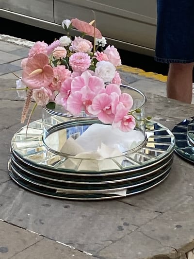 A floral arrangement of pink and white flowers, including anthuriums and orchids, is placed in a clear glass vase. The vase sits on a stack of decorative mirrored plates with faceted edges. The plates are dark-colored with reflective surfaces. In the background, a person wearing a dark blue denim skirt is partially visible, with their legs and lower torso in the frame. The scene appears to be outdoors, possibly on a paved surface or sidewalk, with hints of a building or vehicle in the background. The lighting suggests daylight. The location is identified as Bakikhanov, Azerbaijan.