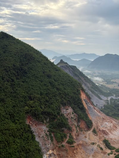 The image shows a panoramic view of a lush green mountain range under a cloudy sky. The foreground is dominated by the steep, tree-covered slope of a mountain, which transitions into a barren, rocky expanse where the mountain has been excavated, revealing layers of reddish-brown and grey soil. A winding dirt road snakes its way through the excavated area, suggesting human activity, possibly related to mining or construction. In the distance, rolling hills and mountains are shrouded in mist, creating a layered effect. Patches of vegetation are visible in the valleys between the distant peaks, and a small village or settlement appears to be nestled within the landscape. The overall scene depicts a blend of natural beauty and human impact on the environment in Hong Ngern Village, Laos.