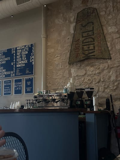 A vintage "Keidel's Soda" sign hangs on a stone wall, next to a coffee shop menu listing various drinks and their prices: coffee, matcha latte, chai latte, hot cocoa, hot tea, iced tea, lemonade, and mimosa. Special teas with flavors like peppermint and holiday chai are also advertised. Below are options like cookie butter latte, peppermint mocha, and white mocha. A row of coffee dispensers sits on a counter.
