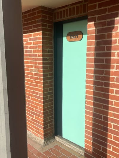 The image displays an architectural detail, specifically a light teal-colored door with a dark frame, marked with the number "201" on an oval-shaped light brown wooden plaque. The door is set within an entryway constructed of reddish-brown bricks with light mortar, arranged in a stretcher bond pattern. A grey concrete pillar is partially visible on the far left, casting a shadow. The ground immediately in front of the door consists of reddish-brown paving bricks. A dark grey, textured threshold or base separates the brick wall from the paved surface. Strong, direct sunlight illuminates the scene, creating distinct shadows, including a prominent vertical shadow on the right brick wall. The upper area reveals a light-colored overhang or ceiling. No individuals, activities, or additional objects are present. This scene is located in the city of Bogota, Capital District, Colombia.