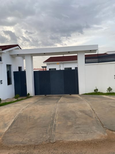 This is a frontal, eye-level photograph of a modern house entrance in Barikin Jirga Summa, Nigeria. The scene is dominated by a large, dark grey metal gate, which is closed. The gate is flanked by white pillars and a white horizontal beam, forming an entryway. To the left of the gate is a section of a white house with a dark-framed window. To the right, a white wall topped with a black security fence extends into the background. The roof of the house is visible above the entryway and along the white wall, featuring a reddish-brown tile. The ground in front of the gate is a concrete driveway, showing signs of wear and cracking, with sparse green grass and small plants growing along the edges. The sky is overcast with heavy, grey clouds.