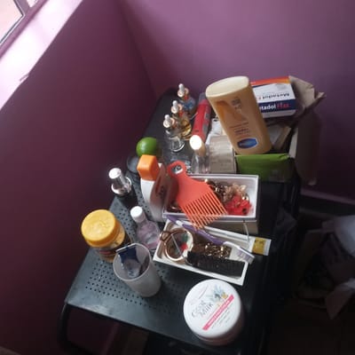 The image displays a collection of personal care products and toiletries neatly arranged on a black, perforated surface, likely a table or trolley. The background features a solid purplish-pink wall, with natural light entering from a window partially visible in the upper left corner, casting a bright stripe on the wall.
Key items visible include several small bottles with droppers, possibly serums, some with visible text like "MOTO SERUM GLOW". A large bottle of Vaseline lotion, labeled "dry skin repair" and "48H moisture", stands upright. Next to it is a box of "Metadol Fizz". An orange hair pick is prominently placed in a white container filled with various hair accessories such as clips and decorative pieces. Below this container, a purple toothbrush and a black brush are visible.
Other products include a white container of "Goat Milk Vitamin E HAND & FACE CREAM" by Doctor White, with Arabic text indicating it's a moisturizing cream for face and body. A white cup holds a tube labeled "Anusol". A yellow-capped jar, a white sunscreen bottle marked "50", and several perfume or oil bottles are also present. A green lime or similar fruit is placed among the bottles.
The scene depicts an organized personal grooming station, possibly in a bedroom or bathroom within a home in Mahiakalo ward, Kenya. No activity is taking place, but the arrangement suggests regular use. The time of day appears to be during daylight hours due to the natural light from the window.