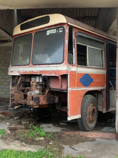 The image shows an abandoned, dilapidated red and cream-colored bus parked in a covered concrete structure. The front of the bus is heavily rusted and damaged, with exposed engine parts. The windows are grimy and one of the front tires appears deflated. A blue circular emblem with a horizontal bar is visible on the side of the bus. The ground is dirty and littered with debris, and weeds are growing around the bus. The structure appears to be a bus garage or workshop. The overall scene suggests disuse and decay. The location is identified as Habaraduwa, Sri Lanka.