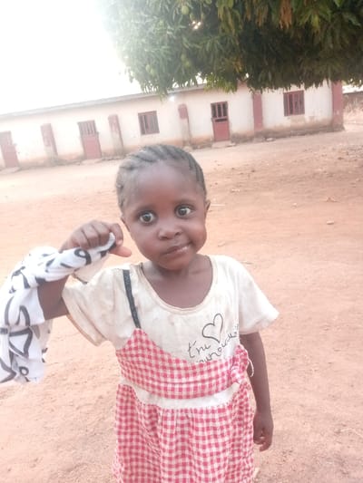 An outdoor scene in Agban, Nigeria, depicts a young child standing on reddish-brown, dusty earth, looking directly at the camera. The child has dark skin and short, dark braided hair. They wear a soiled, light-colored short-sleeved t-shirt with a black heart graphic and partially visible French text ("t'es m'a merveille") over a red and white checkered dress or pinafore, one black strap visible on the left shoulder. The child's left hand is raised, clutching a crumpled white cloth with dark patterns. In the background, a single-story, light-colored building with multiple brown doors and barred windows is visible. Overhanging the building from the upper center is a tree with green foliage and visible fruit. The scene is illuminated by bright daylight, with a bright spot appearing in the upper left of the frame.