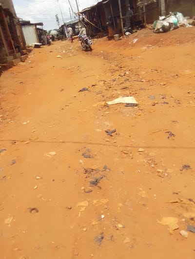 The image depicts an unpaved, reddish-brown dirt road in Nsugbe, Nigeria, under daylight conditions with a partially cloudy sky. The road surface is uneven, littered with small stones, debris, and dark patches of unknown material. A white piece of paper or plastic lies in the mid-foreground.

On the left side of the road, several simple, single-story structures with corrugated metal roofs and walls are visible, appearing to be shops or residences. A white rectangular object is situated in front of one structure. A parked motorcycle with chrome accents is positioned on this side, facing away from the viewer. Further down the road, a person is partially visible near these structures.

On the right side of the road, a longer, weathered building with a dark facade and a low roofline is present, also appearing to be a commercial establishment. Various items, including several large, light-colored sacks (possibly containing goods or refuse), are stacked outside this building. A white bucket is on the ground near the building. An individual wearing light-colored clothing walks away from the camera, further down the road on this side.

Utility poles with overhead power lines span the scene, suggesting an active street or settlement. No explicit interactions are discernible.
