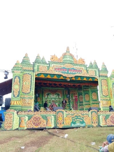 The image shows a stage decorated with a green and gold color scheme. The stage is ornately decorated with gold trim and elaborate patterns, giving it a traditional feel. There are several people on the stage, all appearing to be wearing traditional Indonesian clothing. They are interacting with each other, possibly engaged in a performance. The stage is located in front of a grassy field and is a large, elaborate stage that is likely used for public performances. There is a sign above the stage that reads "Wahyu Budoyo." The stage is located in the city of Kecamatan Margorejo, Indonesia at the coordinates -6.735, 111.016. The image is taken from a ground-level perspective. The sky is clear and there are no clouds visible.