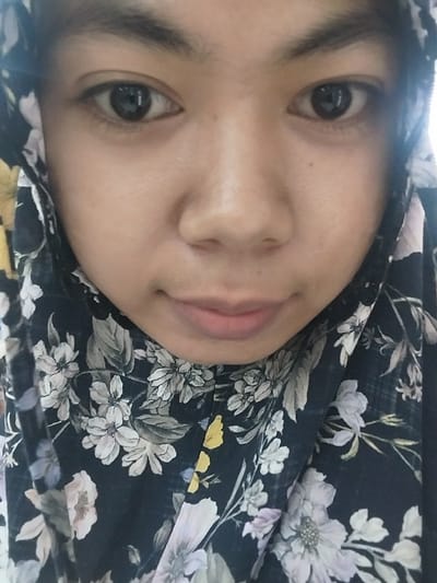 This media file is a close-up, slightly tilted selfie of a young person wearing a dark-colored hijab with a floral pattern. The subject's face is the primary focus, with one eye and part of their mouth visible. The background is dominated by the patterned fabric of the hijab, which features light-colored flowers, possibly in shades of white, pink, and yellow, against a dark background. The lighting appears to be natural, suggesting the photo was taken during the day. The subject's expression is neutral. There is no visible text in the image. The setting appears to be indoors or in a location where the subject is wearing the hijab. No specific time of day or weather conditions can be determined from the image.