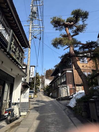 This image depicts a narrow, uphill street in Nozawaonsen, Japan, under a clear blue sky. The scene is devoid of people.
On the left, a two-story building with a dark tiled roof and white lower facade features a large, weathered wooden sign displaying Japanese characters, reading "松葉屋" (Matsubaya). A cream-colored motorcycle with a rear rack and a visible license plate number "10" is parked on the sidewalk in front of this building. Directly above, a utility pole densely packed with transformers, cross arms, and numerous overhead power lines dominates the upper left quadrant. Further up the street on the left, another vertical sign reads "居酒屋 は車" (Izakaya Hakuruma).
On the right side of the street, a large, gnarled pine tree with twisted branches and green needles stands prominently, leaning towards the street. Behind it, a two-story building features dark brown wooden siding on its upper level, a stone-clad base, and a traditional street lamp affixed to its facade. Patches of snow are visible along the base of the stone wall and tree on this side. Further along the street on the right, another building with lighter wooden siding is visible.
The paved street ascends gently uphill, curving slightly to the left in the distance. The background reveals additional traditional-style buildings and forested hillsides under the bright daylight. No specific actions or interactions are visible.