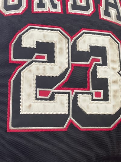 This media file features a close-up view of a dark-colored garment, likely a sports jersey, adorned with large, multi-layered embroidered numbers and text. No people or activities are depicted; the image focuses entirely on the garment's details.
The central elements are the large numbers "33". To the left of these numbers, partially visible embroidered letters spell out the word "SPORTS". The garment itself is made of a dark, tightly woven fabric, possibly black or a very dark navy blue.
The numbers and letters are constructed with a layered, appliquéd technique. The innermost layer is a vibrant red fabric. This is framed by a layer of the dark garment fabric, followed by an outermost layer of off-white material. The edges of the off-white layer are defined by tight, zigzag stitching in white thread, giving it a textured border. Red stitching also outlines the red portions of the numbers.
A notable detail is the condition of the off-white layers, which display significant discoloration, appearing dingy with various brownish-grey stains and smudges, suggesting extensive wear or age. The overall aesthetic is that of a well-used piece of athletic attire. There are no visual cues within the image to indicate the time of day, weather, or a specific location in Troyan, Bulgaria, as only the textile surface is shown.
