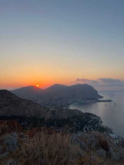 The image captures a panoramic view of the Mondello coast near Palermo, Italy, during a sunset. The sun, a bright orange orb, is positioned just above the silhouette of a mountain range, casting a warm glow across the sky. The sky transitions from a soft orange at the horizon to a pale blue overhead.
In the foreground, rough, rocky terrain with dry, golden grasses and shrubs dominates the lower portion of the frame, suggesting a vantage point from a hillside.
Further in the distance, a crescent-shaped bay is visible, lined with a dense collection of white buildings that form the town of Mondello. The architecture suggests a Mediterranean style, with houses clustered closely together. The bay is dotted with several boats, including larger vessels that appear to be ships or ferries, and smaller sailboats.
The sea is calm, reflecting the muted colors of the sky. Mountains with rugged, dark slopes rise behind the town, some of them covered in sparse vegetation. The overall atmosphere is serene and picturesque, typical of a coastal sunset. No people are discernible in the image.