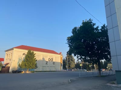 The image depicts a street scene in Antratsyt, Ukraine, bathed in the warm light of a setting or rising sun. A two-story building with a red roof dominates the left side of the frame, featuring a large mural of a young man in a denim jacket on its facade. The building's walls are light-colored, with several windows and what appear to be air conditioning units. The words "od ze" are prominently displayed in large, dark letters on the building, with smaller text below that reads "ОДЕЖДА ОБУВЬ АКСЕССУАРЫ" (Clothing, Footwear, Accessories). A dark-colored sedan is parked on the street in front of the building, near the mural.

In the foreground, a paved street runs across the width of the image, with a grey sidewalk and a metal fence separating it from a grassy area. Several evergreen trees are planted in front of the building and along the street. On the right side of the image, part of a modern building with a white, segmented facade is visible, along with a large deciduous tree whose branches extend into the upper portion of the frame. The sky is a clear, bright blue, with a few small birds visible near the top left. Traffic lights and pedestrian crossing signs are present in the background, suggesting an urban environment.