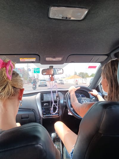 A view from the back seat of a car moving through Hua Hin City Municipality, Thailand. Two women are visible, one driving and the other as a passenger. The driver has visible arm tattoos. The car's interior is predominantly black. Through the windshield, a street scene is visible with other vehicles, buildings, and road signs.