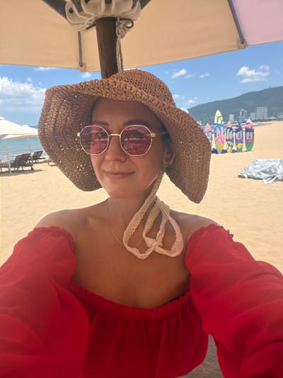 A woman is taking a selfie on a beach in Quy Nhon Ward, Vietnam. She is wearing a straw hat with a wide brim and pink-tinted sunglasses. Her off-the-shoulder red top has puffy sleeves. A light-colored lace detail is visible around her neck and chest. In the background, there is a sandy beach, lounge chairs under umbrellas, and a line of colorful surfboards in front of buildings. The sky is blue with scattered clouds, and a mountain range is visible in the distance.