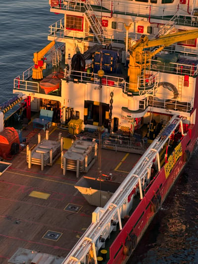 This media file showcases a detailed view of the deck of a large vessel, likely a ship or a platform, seen from an elevated perspective. The scene is bathed in the warm, golden light of either sunrise or sunset, casting long shadows and highlighting the textures of the metal and wooden surfaces.
The deck is densely packed with various equipment, including cranes, winches, containers, and what appear to be storage tanks. There are also safety features visible, such as a "RESCUE ZONE" marked in yellow and red, and fire extinguishers. Ropes and chains are coiled or secured, indicating an active operational environment. A few people in work attire are visible, engaged in tasks or observing the surroundings.
The vessel itself has a white and red superstructure, with numerous windows and access points. The surrounding water is calm and a deep blue, reflecting the colors of the sky and the vessel. The overall impression is one of industrial activity and maritime operations, captured during a serene moment of the day. Text visible in the image includes "RESCUE ZONE," "MUD TK. 2," "MUD TK. 3," "COLOR CODE," and "SWL 2t-15m," providing specific operational details about the equipment and areas on the vessel.