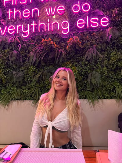 This is a portrait-oriented, medium shot photograph of a young woman in an indoor setting, possibly a restaurant or bar, in Patong, Thailand. The woman has long blonde hair with some pink highlights. She is smiling and looking towards the camera. She is wearing a white tied top and plaid shorts. Behind her, there is a vertical wall covered in artificial greenery, resembling a living wall. To the left of the woman, a bright pink neon sign is mounted on the green wall. The neon sign displays text in a vertical orientation. The visible text reads, "If then we do everything else." The overall mood of the image is cheerful and vibrant, with the neon lights and the woman's smile contributing to a positive atmosphere. The lighting appears to be artificial, likely from the neon sign and ambient room lights, and it casts a warm glow on the scene. The background suggests an evening or indoor setting where artificial lighting is used.