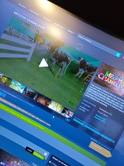 The image shows a computer screen displaying the Steam store page for a game called "MECCHA CHAMELEON". The screen is angled and partially illuminated by an overhead light source, creating glare. The game's title is prominently displayed in large, colorful letters. A description of the game is visible, stating: "Paint yourself to blend in! "Meccha Chameleon" is a sensation hide-and-seek game where you paint your white body to mimic the stage. Spot, pose, and skill" are keys to survival. Deceive Seekers with techniques that put chameleons to shame! Supports public matches and streaming." Below this, there are details about the game including release date, developer, and publisher. A row of smaller thumbnail images, presumably screenshots or videos from the game, are visible along the bottom of the screen. A green bar with text in Russian reads: "MECHCHA CHAMELEON already in your Steam library." There is also a price displayed as "1 930T" and a button for "Add to cart". The background of the Steam interface is a dark blue. The screen itself appears to be a laptop or desktop monitor. There is no information in the image that provides any context or indication of the location being in the city of Оқжетпес, Kazakhstan, or any other specific geographic location.