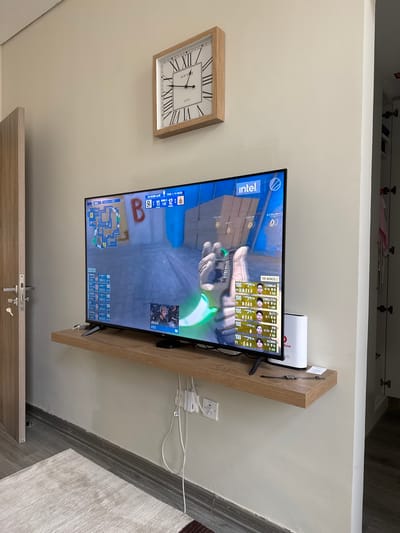 In Doha, Qatar, a living room features a wall-mounted television displaying a first-person shooter video game, likely Counter-Strike, with a prominent "intel" logo in the upper right corner. The game interface shows player statistics and a minimap. To the left of the television, a wooden shelf holds a white modem or router. Above the TV, a square clock with Roman numerals hangs on the wall. A light-colored door with a metallic handle is visible on the far left, and a closet door with hinges is on the right. A beige rug covers part of the dark wooden floor in the foreground, and white cables are neatly managed along the wall with clips.