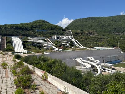 An abandoned aquapark in Podostrog, Montenegro, is depicted under a clear blue sky with scattered white clouds. Lush green hills form the backdrop to the scene. The aquapark features numerous white water slides, some of which are elaborate, spiraling structures, while others are wide, straight drops. The slides are integrated into the sloped terrain, with metal support structures visible.
Various pools are present; some appear empty and disused, while others contain stagnant, murky green water. The surrounding area is overgrown with wild vegetation, with bushes and weeds encroaching on the paved walkways and structures. Some of the paving stones are cracked and uneven, with plants growing through the gaps. A concrete wall runs along a section of the aquapark, showing signs of wear and tear. There are no people visible in the image. The overall impression is one of decay and neglect.