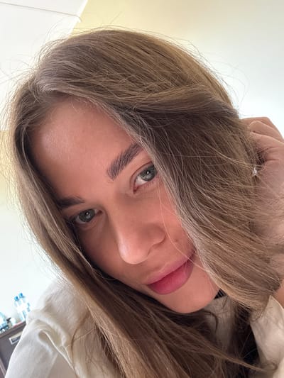 A close-up, slightly angled downward selfie of a woman with light brown hair partially obscuring her face. Her eyes are grey-blue, and she is wearing pink lipstick. The background appears to be a neutral off-white color, possibly a wall. A white garment is partially visible in the lower right corner. There is no evidence to support a location in Sosnovy, Russia.
