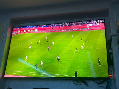 A television screen shows a soccer game between Algeria (ALG) and Nigeria (NGA). The score is 0-2 in favor of Nigeria at 77:39 minutes into the match. The game is being broadcast on beIN SPORTS 1. The pitch is green with white markings, and players in white and green jerseys are visible. The field is surrounded by spectators. Multiple "Max it" banners are also visible. A man in a black suit stands near the bottom of the screen.