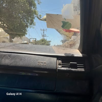 The image shows the interior of a vehicle, looking out through the windshield. The dashboard is visible in the foreground, with the "SRS AIRBAG" text embossed on it. Through the windshield, the exterior environment of Deir el-Balah, Palestinian Territories, is visible. There is a large green tree to the left, and buildings and utility poles in the background. A large cloth banner with an apple logo is draped over something in the distance, partially obscuring buildings and market stalls. The sky is clear and blue. The image is dated as captured by a "Galaxy A14".