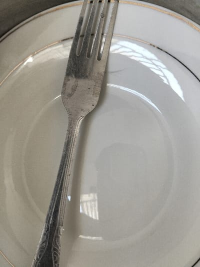 A top-down close-up shows a silver fork resting on a white plate with a gold rim. The fork's tines and handle are visible, with slight tarnish. The plate's surface reflects light, creating bright areas. The plate's center is largely empty.