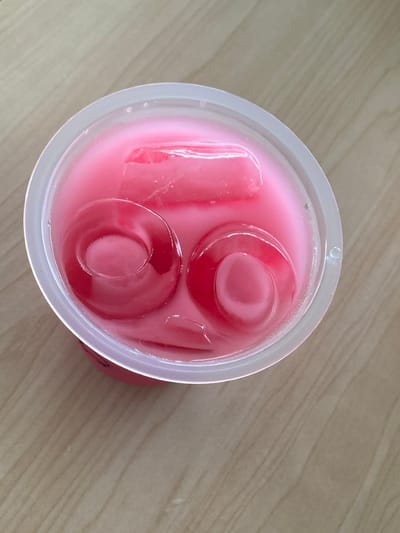 A top-down shot captures a vibrant pink drink served in a clear, disposable plastic cup, resting on a light-colored wooden surface with a visible grain pattern. The cup contains an opaque pink liquid and several ice cubes. Some ice cubes are rectangular, while others exhibit a distinct cylindrical or ring-like shape. The plastic cup shows slight condensation or frost on its exterior, indicating the beverage is cold. The scene depicts a casual setting, likely indoors, in Bekasi Regency, Indonesia. No people or visible text are present in the image. The time of day and weather are not discernible.