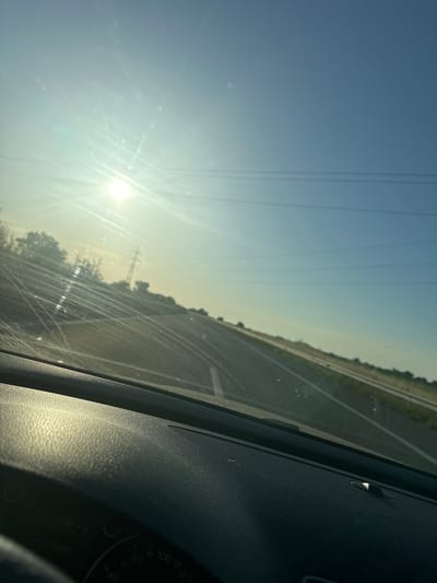 The image is taken from inside a moving car on a road near Shishmantsi, Bulgaria. The sun is brightly shining in the sky, with power lines visible in the distance. Part of the car's dashboard and a section of the speedometer are visible in the lower right of the frame. The windshield is scratched and slightly dirty. The road appears to be a highway. Trees and vegetation line the side of the road.